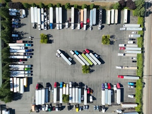 An aerial view of a parking lot filled with trucks and trailers arranged in rows. The vehicles are organized in a systematic manner, with some smaller cars and trailers mixed in. Trees and small green spaces are distributed throughout the area, providing some greenery amid the concrete.