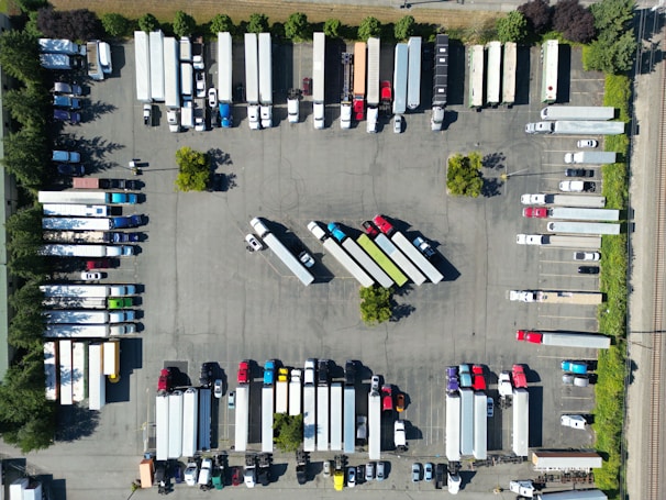 An aerial view of a parking lot filled with trucks and trailers arranged in rows. The vehicles are organized in a systematic manner, with some smaller cars and trailers mixed in. Trees and small green spaces are distributed throughout the area, providing some greenery amid the concrete.