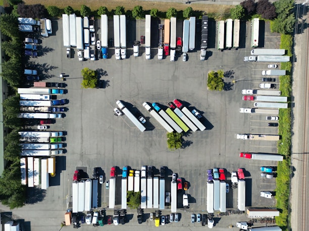 An aerial view of a parking lot filled with trucks and trailers arranged in rows. The vehicles are organized in a systematic manner, with some smaller cars and trailers mixed in. Trees and small green spaces are distributed throughout the area, providing some greenery amid the concrete.