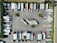 An aerial view of a parking lot filled with trucks and trailers arranged in rows. The vehicles are organized in a systematic manner, with some smaller cars and trailers mixed in. Trees and small green spaces are distributed throughout the area, providing some greenery amid the concrete.