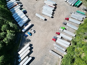 Aerial view of a parking area filled with several semi-trailers, arranged in neat rows. The trailers are primarily white, with a few in different colors such as red, blue, and green. The area is surrounded by greenery, with trees lining the lot on one side.