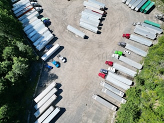 Aerial view of a parking area filled with several semi-trailers, arranged in neat rows. The trailers are primarily white, with a few in different colors such as red, blue, and green. The area is surrounded by greenery, with trees lining the lot on one side.