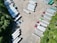 Aerial view of a parking area filled with several semi-trailers, arranged in neat rows. The trailers are primarily white, with a few in different colors such as red, blue, and green. The area is surrounded by greenery, with trees lining the lot on one side.
