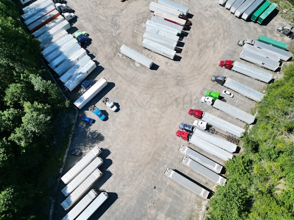 Aerial view of a parking area filled with several semi-trailers, arranged in neat rows. The trailers are primarily white, with a few in different colors such as red, blue, and green. The area is surrounded by greenery, with trees lining the lot on one side.