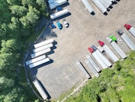 An aerial view of a parking lot with multiple semi-trailers and a few trucks parked in different orientations. The area is surrounded by dense green trees, providing a natural border. The ground is asphalt, and there are noticeable shadows cast by the vehicles and trees.