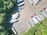 An aerial view of a parking lot with multiple semi-trailers and a few trucks parked in different orientations. The area is surrounded by dense green trees, providing a natural border. The ground is asphalt, and there are noticeable shadows cast by the vehicles and trees.