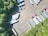 An aerial view of a parking lot with multiple semi-trailers and a few trucks parked in different orientations. The area is surrounded by dense green trees, providing a natural border. The ground is asphalt, and there are noticeable shadows cast by the vehicles and trees.