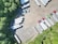 An aerial view of a parking lot with multiple semi-trailers and a few trucks parked in different orientations. The area is surrounded by dense green trees, providing a natural border. The ground is asphalt, and there are noticeable shadows cast by the vehicles and trees.