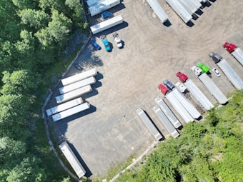 An aerial view of a parking lot with multiple semi-trailers and a few trucks parked in different orientations. The area is surrounded by dense green trees, providing a natural border. The ground is asphalt, and there are noticeable shadows cast by the vehicles and trees.