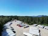 A large, open parking area filled with numerous semi-truck trailers is surrounded by dense, green forest. The sky is clear and blue with mountains visible in the background. Several red and yellow trucks are parked among the rows of white trailers.
