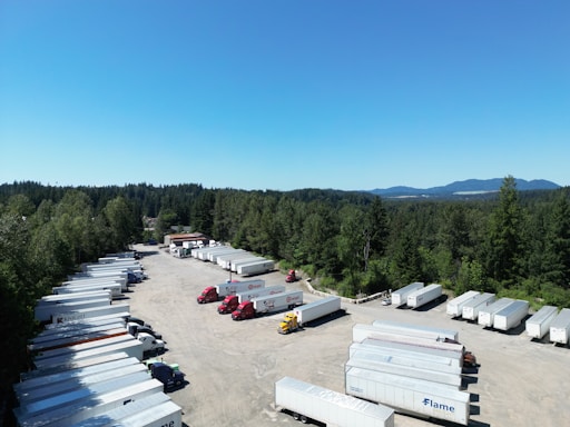 A large, open parking area filled with numerous semi-truck trailers is surrounded by dense, green forest. The sky is clear and blue with mountains visible in the background. Several red and yellow trucks are parked among the rows of white trailers.