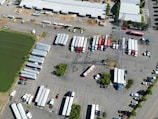 An aerial view of a large parking area filled with numerous semi-trailers and trucks arranged in rows. A prominent electrical transmission tower stands in the middle of the parking lot. Buildings with white roofs are visible at the top of the image, and a green water body is seen on the left. The scene is well-organized, with vehicles lined up neatly, and only a few cars are scattered around.