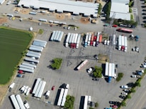 An aerial view of a large parking area filled with numerous semi-trailers and trucks arranged in rows. A prominent electrical transmission tower stands in the middle of the parking lot. Buildings with white roofs are visible at the top of the image, and a green water body is seen on the left. The scene is well-organized, with vehicles lined up neatly, and only a few cars are scattered around.