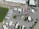 An aerial view of a large parking area filled with numerous semi-trailers and trucks arranged in rows. A prominent electrical transmission tower stands in the middle of the parking lot. Buildings with white roofs are visible at the top of the image, and a green water body is seen on the left. The scene is well-organized, with vehicles lined up neatly, and only a few cars are scattered around.