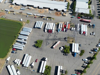 An aerial view of a large parking area filled with numerous semi-trailers and trucks arranged in rows. A prominent electrical transmission tower stands in the middle of the parking lot. Buildings with white roofs are visible at the top of the image, and a green water body is seen on the left. The scene is well-organized, with vehicles lined up neatly, and only a few cars are scattered around.