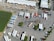 An aerial view of a large parking area filled with numerous semi-trailers and trucks arranged in rows. A prominent electrical transmission tower stands in the middle of the parking lot. Buildings with white roofs are visible at the top of the image, and a green water body is seen on the left. The scene is well-organized, with vehicles lined up neatly, and only a few cars are scattered around.