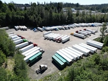 Aerial view of a large parking area filled with numerous trucks and trailers. The area is surrounded by dense greenery with tall trees. Various trailers are parked in organized rows, and there is a mixture of truck cabs and trailers.