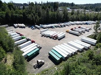 Aerial view of a large parking area filled with numerous trucks and trailers. The area is surrounded by dense greenery with tall trees. Various trailers are parked in organized rows, and there is a mixture of truck cabs and trailers.