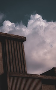 A modern architectural structure with vertical slats stands against a backdrop of large, billowing clouds in the sky. The building has a concrete appearance, with dark, muted shades creating a stark contrast with the lighter, fluffy clouds.