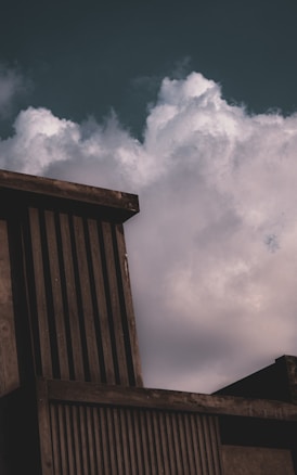 A modern architectural structure with vertical slats stands against a backdrop of large, billowing clouds in the sky. The building has a concrete appearance, with dark, muted shades creating a stark contrast with the lighter, fluffy clouds.