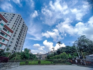 A residential urban scene featuring a tall, multi-story white building with red accents on the left. To the right, a fenced area encompasses lush green trees and grass. The sky is bright and vivid, filled with expansive white clouds against a vibrant blue backdrop.