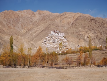 A monastery is nestled against a rugged, brown mountain backdrop under a clear blue sky. Tall trees with autumn foliage surround the base of the mountain, adding shades of yellow and orange to the scenery.