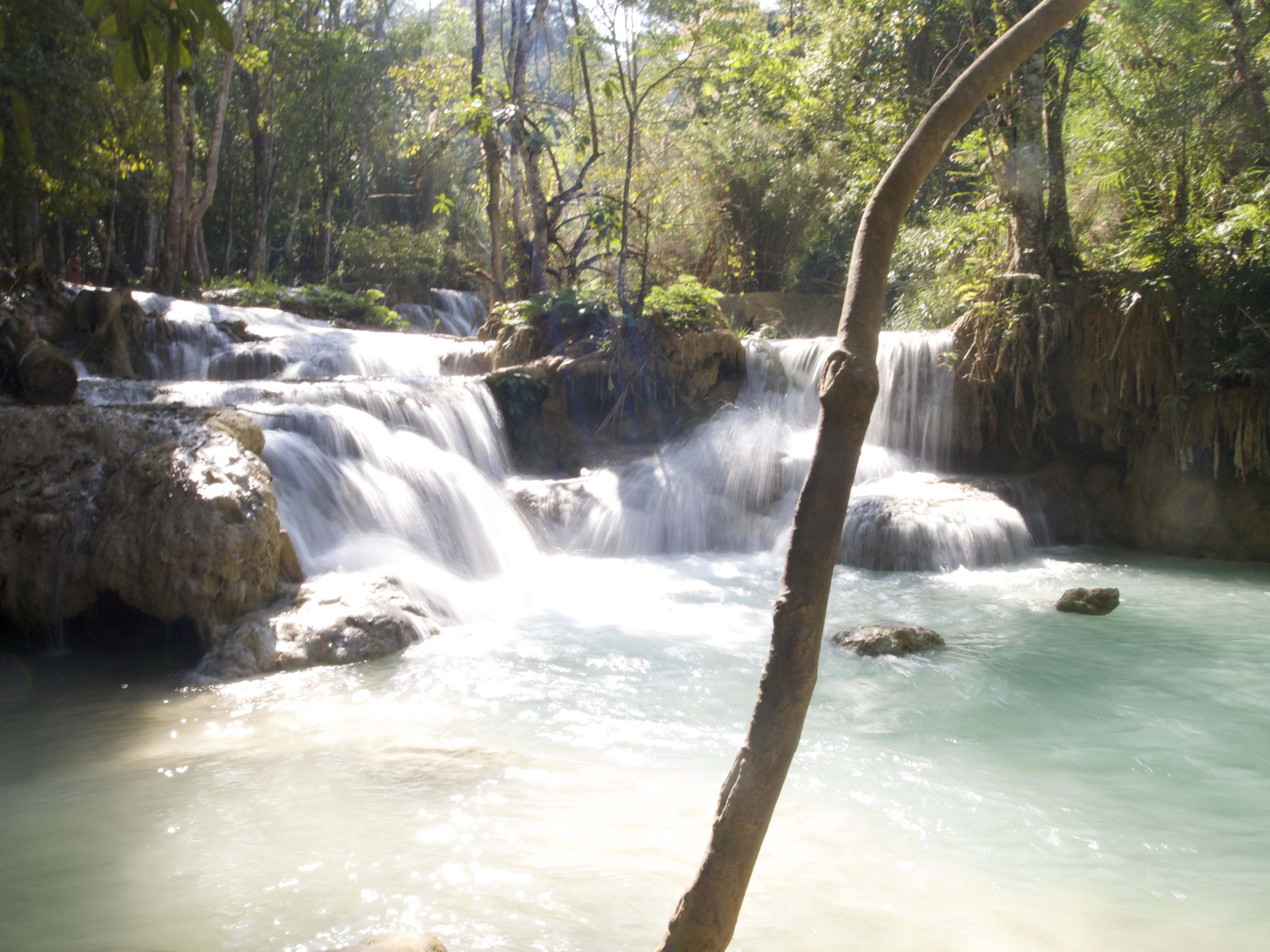 Sunlight filters through trees onto the turquoise cascades and pools of a tiered waterfall.