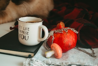 A cozy arrangement featuring a white mug with the phrase 'Let life surprise you', a red pomegranate adorned with a silver chain, and a small pink mushroom. These items rest on a black notebook and a fringed cloth, with a warm red and black plaid blanket in the background. There are hints of a cat's paw in the corner.