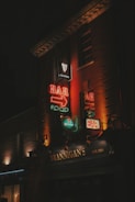 Neon signs illuminate the facade of a building with the name 'Brannigan's' and an emphasis on bar and food. The signs are vibrant with a mix of red, orange, and green lights, advertising brands like Guinness and Heineken. The dark surroundings create a contrast that brings focus to the colorful signage.