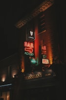 Neon signs illuminate the facade of a building with the name 'Brannigan's' and an emphasis on bar and food. The signs are vibrant with a mix of red, orange, and green lights, advertising brands like Guinness and Heineken. The dark surroundings create a contrast that brings focus to the colorful signage.