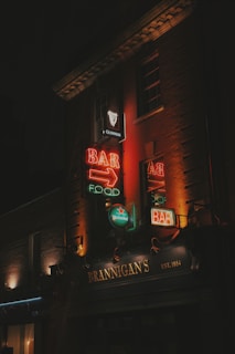 Neon signs illuminate the facade of a building with the name 'Brannigan's' and an emphasis on bar and food. The signs are vibrant with a mix of red, orange, and green lights, advertising brands like Guinness and Heineken. The dark surroundings create a contrast that brings focus to the colorful signage.