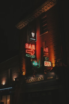 Neon signs illuminate the facade of a building with the name 'Brannigan's' and an emphasis on bar and food. The signs are vibrant with a mix of red, orange, and green lights, advertising brands like Guinness and Heineken. The dark surroundings create a contrast that brings focus to the colorful signage.
