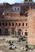 Ancient ruins featuring brick walls with multiple arches and a central column. The background includes old buildings with weathered facades. The ground is scattered with large stone slabs and debris, indicative of historical architecture.
