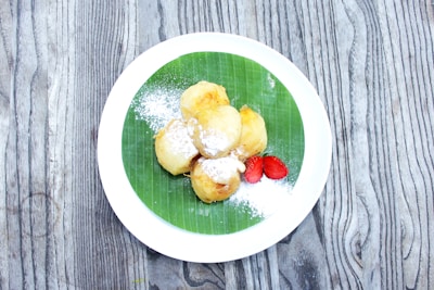 A white plate on a wooden surface holds a few golden brown fried snacks, dusted with powdered sugar, and a couple of sliced red strawberries. The plate features a green banana leaf as a base.