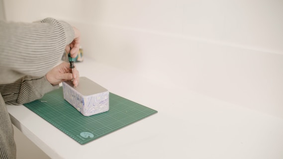 Someone wearing a striped sweater is using a screwdriver to fix or assemble a rectangular object with light blue and purple patterns on its side. The object is placed on a green cutting mat, situated on a white surface or table.