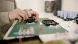 Hands are assembling electronic components on a green cutting mat. Several cylindrical battery cells are visible in the background, along with other electronic parts.