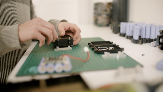 Hands are assembling electronic components on a green cutting mat. Several cylindrical battery cells are visible in the background, along with other electronic parts.