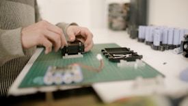 Hands are assembling electronic components on a green cutting mat. Several cylindrical battery cells are visible in the background, along with other electronic parts.