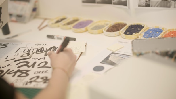 A workspace desk with a person writing or drawing on sheets of paper featuring black calligraphy-style numbers and letters. Several trays containing colorful sequins or beads are lined up in the background, and various stationery items like a marker and paper sheets are scattered around.