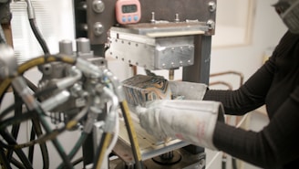 A person wearing protective gloves operates a machine press or hydraulic press, with various knobs, levers, and pipes visible. A patterned block is inside the press, and there is a small digital timer attached to the machine.