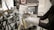 A person wearing protective gloves operates a machine press or hydraulic press, with various knobs, levers, and pipes visible. A patterned block is inside the press, and there is a small digital timer attached to the machine.