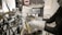 A person wearing protective gloves operates a machine press or hydraulic press, with various knobs, levers, and pipes visible. A patterned block is inside the press, and there is a small digital timer attached to the machine.