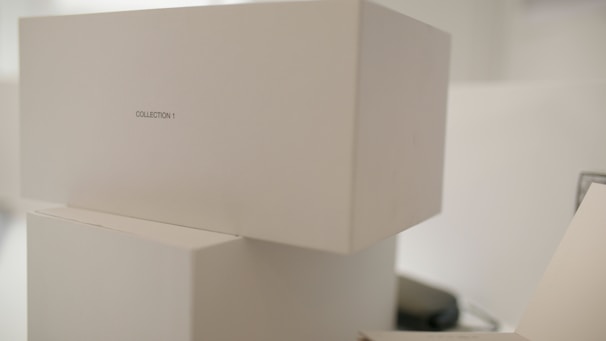 Stacked white boxes with minimalistic design featuring a label 'COLLECTION 1'. The setting appears to be clean and well-organized, possibly in a storage or retail environment.