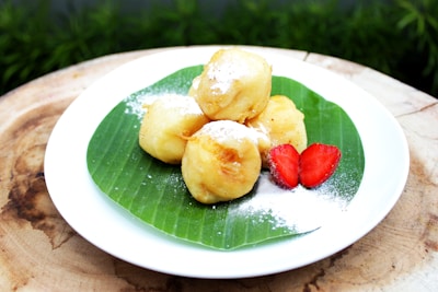 Golden, fried dough balls are arranged on a banana leaf, placed on a white plate. They are dusted with a light layer of powdered sugar. A couple of fresh strawberry slices are added for garnish, providing a pop of red color.