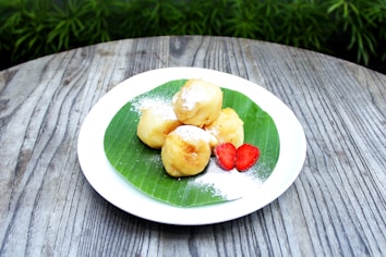 A plate with four fried dough balls topped with powdered sugar is placed on a wooden table. The fried dough balls rest on a green leaf, and there are two slices of strawberry next to them.