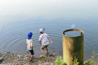 Two children are standing at the edge of a calm body of water, appearing to play or explore. One child is wearing a blue hat and striped shirt, while the other wears a cap and shorts. Nearby, a tall cylindrical metal structure stands partially submerged at the water's edge, with greenery growing around it.
