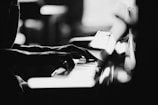 A black and white image featuring a close-up of hands playing a piano. The lighting creates a dramatic and artistic contrast, highlighting the fingers on the keys with a soft out-of-focus background.