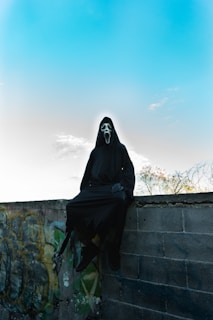 A person wearing a black cloak and a white ghost mask sits on a graffiti-covered wall under a bright blue sky. The overall atmosphere feels eerie and mysterious with elements of urban decay and open sky.