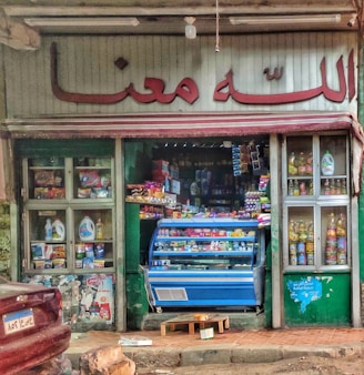 A small, colorful storefront selling groceries and household items. The entrance is filled with various products, including groceries in a blue display fridge. Shelves on either side showcase items like cooking oils and detergents. The store has a sign in a language that uses an Arabic script above the door.
