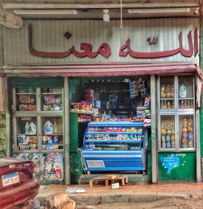 A small, colorful storefront selling groceries and household items. The entrance is filled with various products, including groceries in a blue display fridge. Shelves on either side showcase items like cooking oils and detergents. The store has a sign in a language that uses an Arabic script above the door.